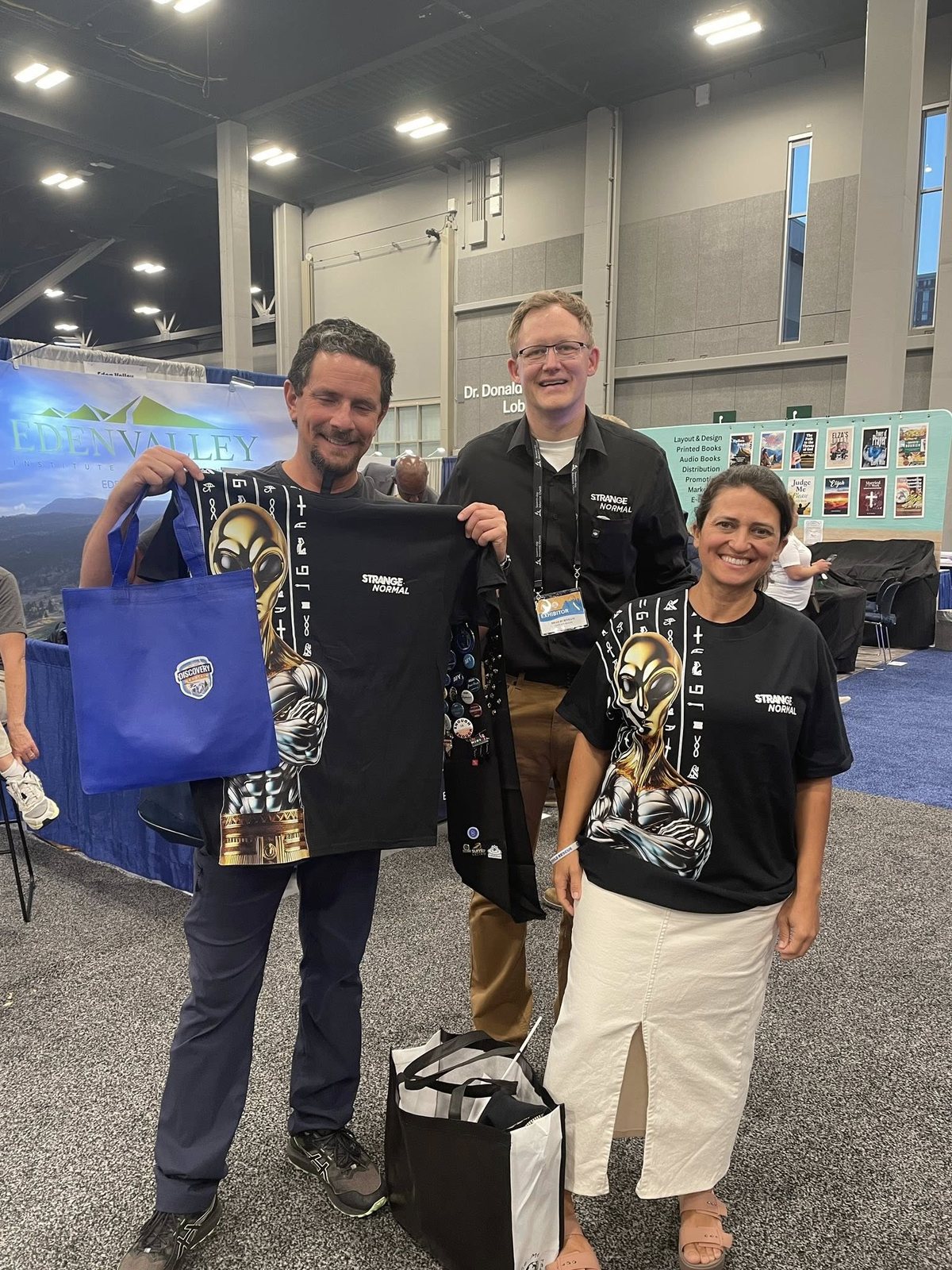 Three Strange Normal team members holding conference tote bags at an exhibition stand