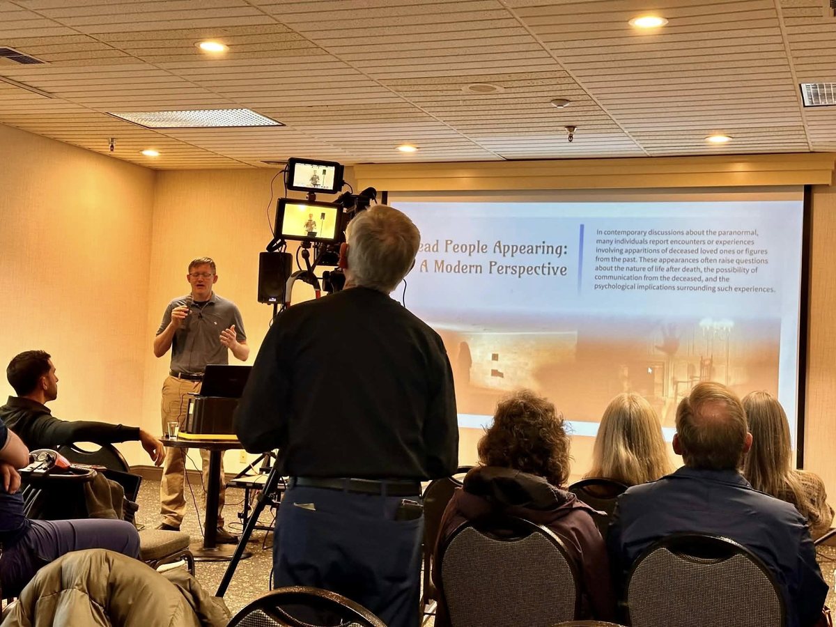 A presentation in progress before a seated congregation, with a projection screen and camera rig