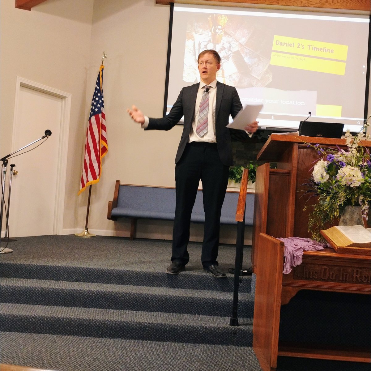 Bradley Burnham speaking at a podium beside an American flag, with a slide projected behind him